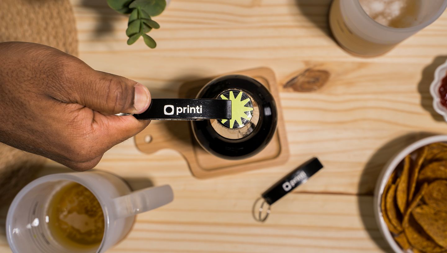 Pessoa utilizando um Chaveiro Abridor Clane personalizado da Printi, em cima de uma mesa de madeira e com canecas ao redor