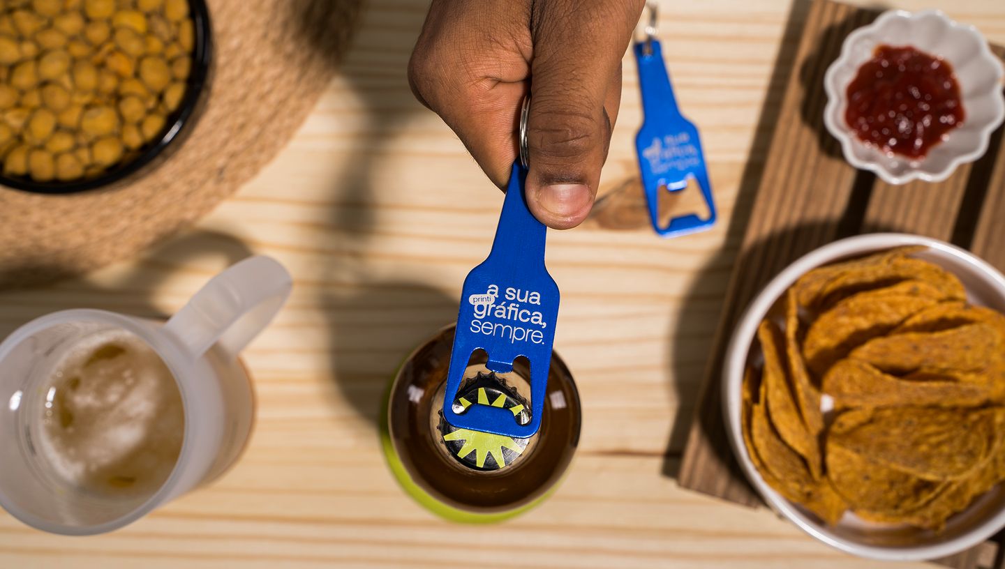 Mão de uma pessoa utilizando um chaveiro alumínio abridor garrafa personalizado na cor azul para abrir uma garrafa de bebida em uma mesa de madeira, cercada por petiscos, molho e copo de cerveja
