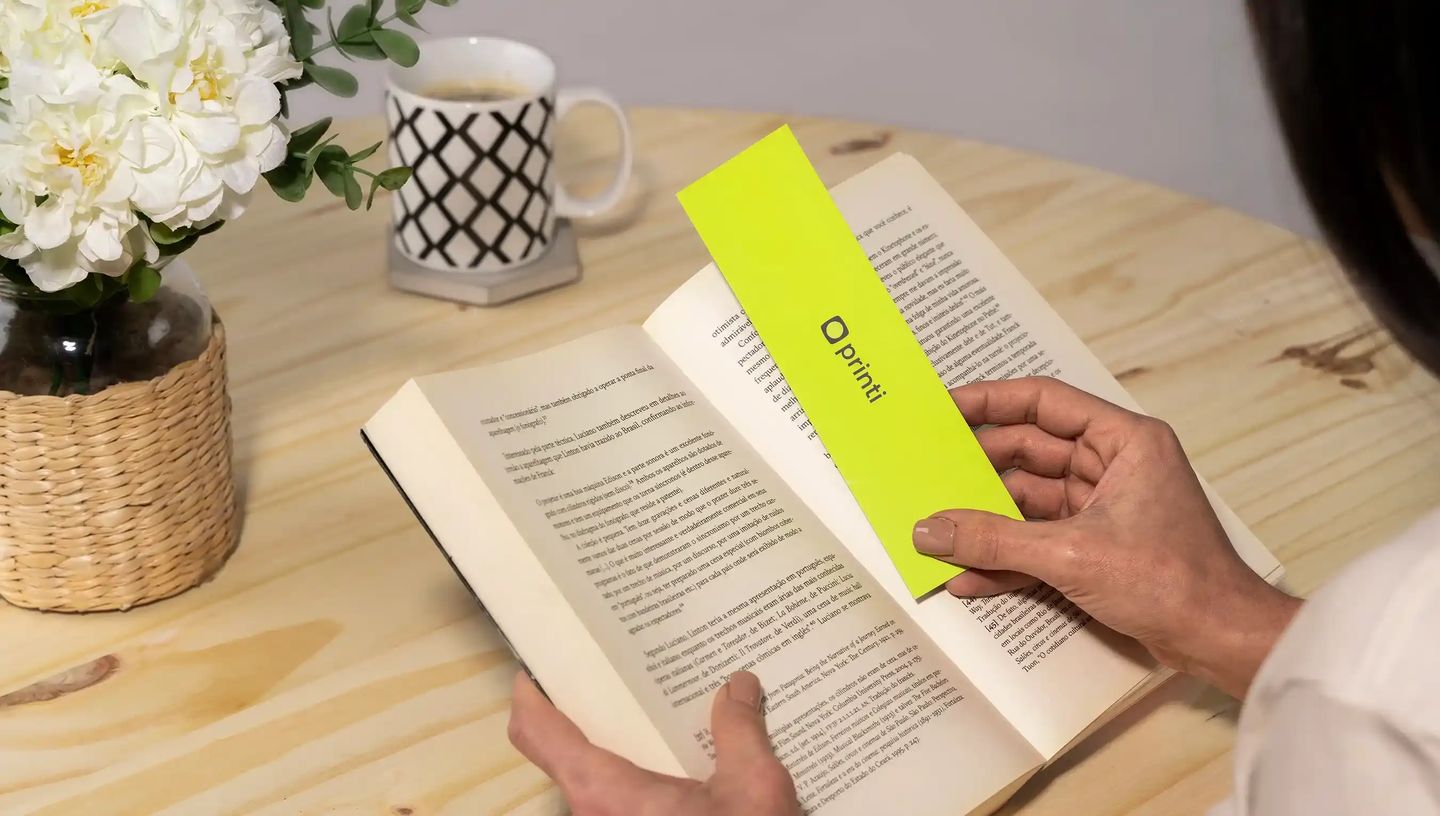 Marca-página personalizado verde neon com logotipo Printi sendo utilizado em livro aberto.