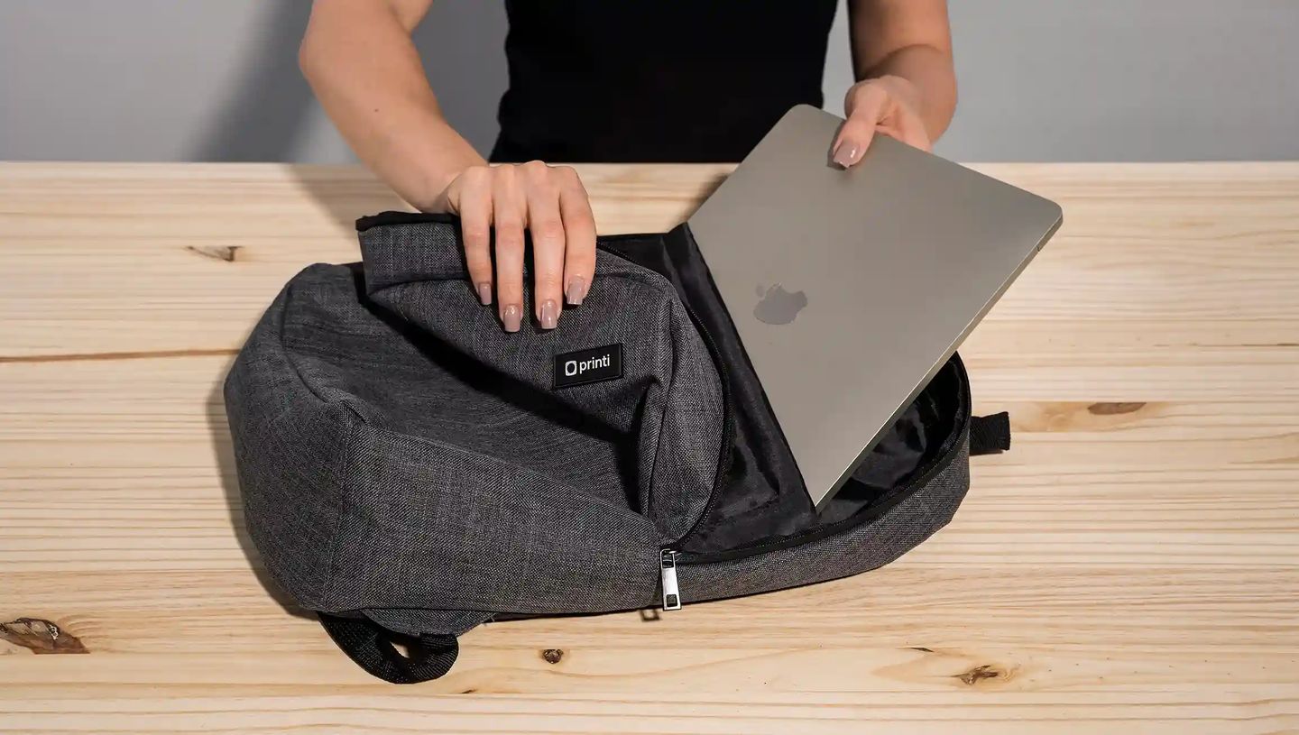 Mochila personalizada para notebook da Printi aberta sobre mesa de madeira enquanto uma pessoa coloca um laptop dentro do compartimento
