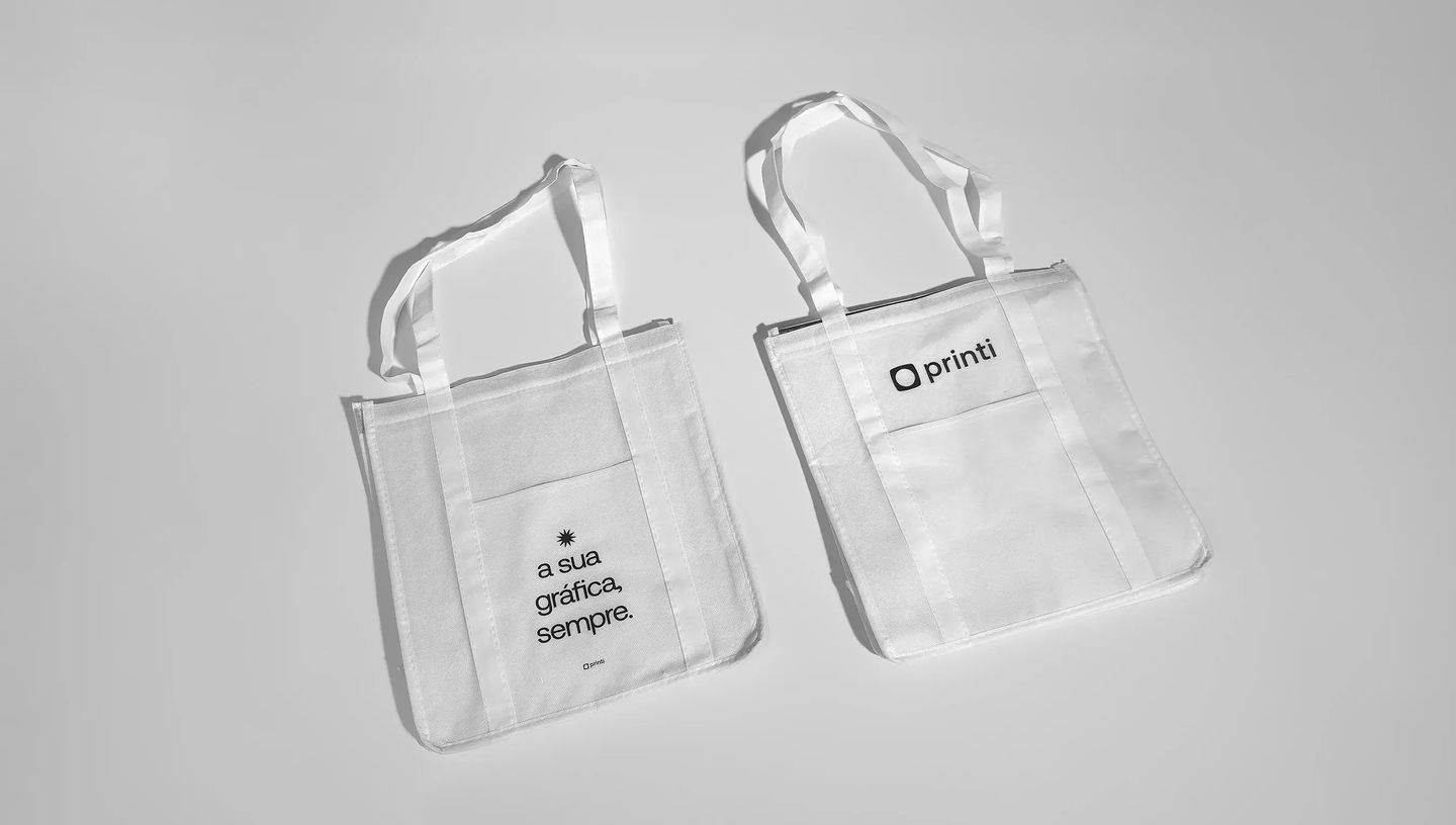 Duas sacolas térmicas personalizadas de tecido branco com alças longas, posicionadas lado a lado sobre fundo neutro. Uma delas exibe a frase “a sua gráfica, sempre.” e a outra apresenta o logo da Printi na parte superior.