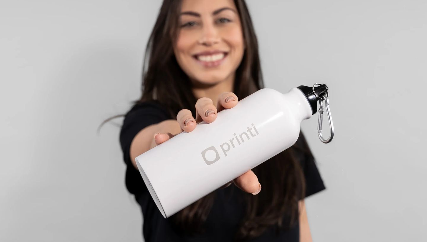 Mulher sorrindo segura uma squeeze de alumínio personalizada branca com logotipo da Printi. A garrafa possui tampa preta com mosquetão metálico e está sendo apresentada em primeiro plano, destacando o produto promocional personalizado.