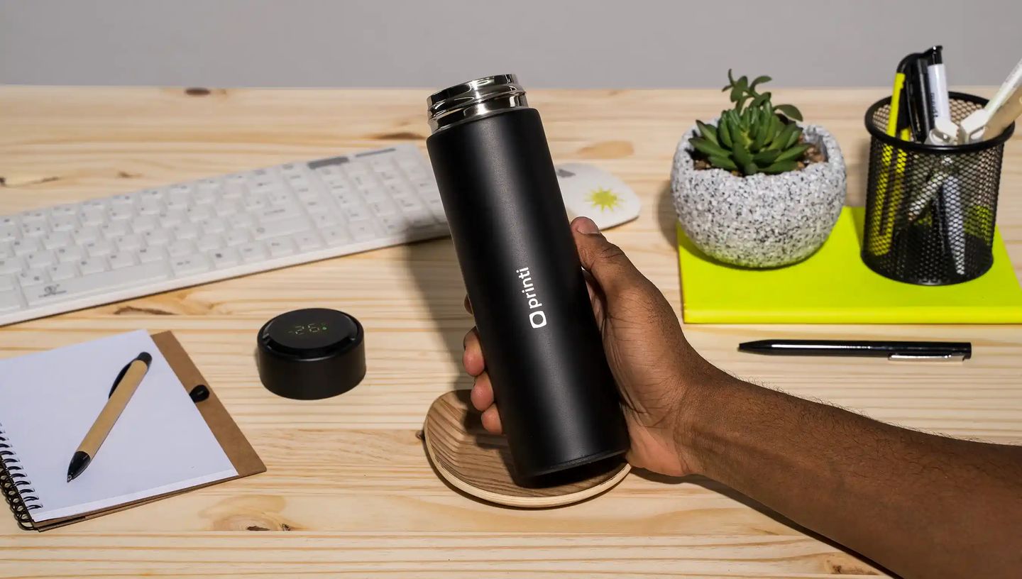 Pessoa segurando squeeze térmico personalizado da Printi em aço inox com acabamento preto fosco sobre mesa de madeira em ambiente de escritório. Ao redor aparecem teclado, caderno, caneta, porta-lápis e planta decorativa