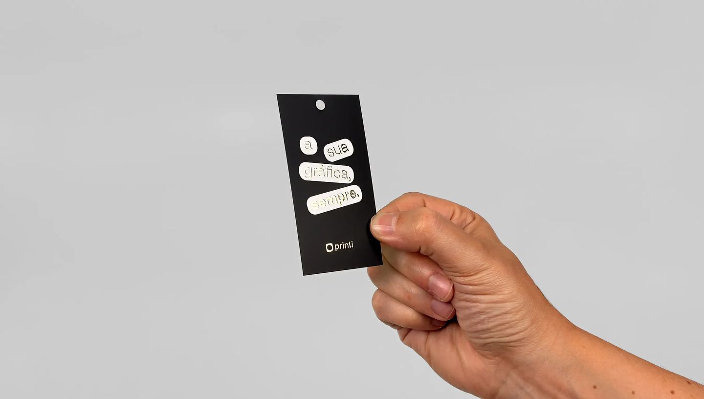 Mão segurando tag personalizada preta na vertical, com furo superior e aplicação em relevo branco com a frase “a sua gráfica, sempre”, sobre fundo cinza claro.