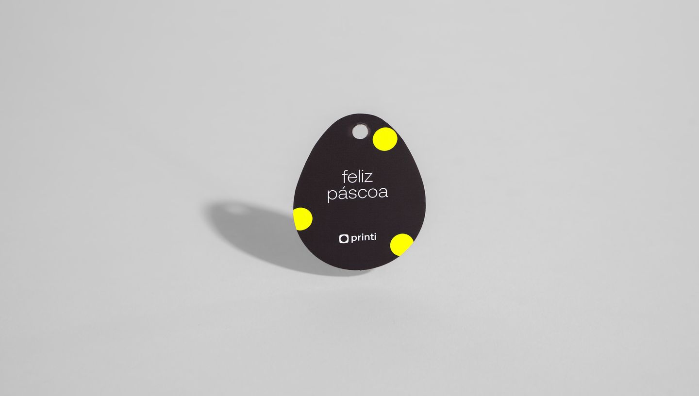 Tag para Ovo de Páscoa individual no formato de ovo, com fundo preto. O design inclui bolinhas amarelas posicionadas nos cantos e o texto “feliz páscoa” em branco centralizado, acompanhado da marca Printi na parte inferior.
