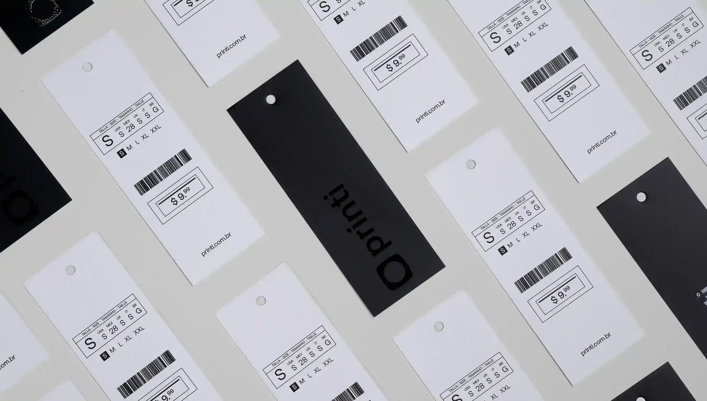 Conjunto de tags para roupas personalizadas dispostas sobre fundo claro, com etiquetas brancas exibindo informações de tamanho, código de barras e preço, além de tags pretas com o logo “printi”, ilustrando etiquetas de vestuário usadas para identificação 