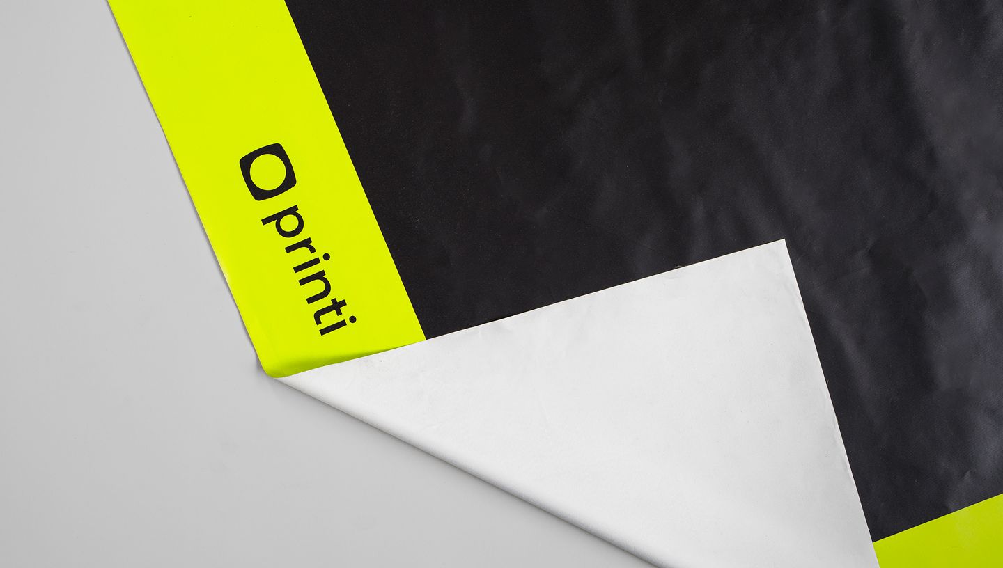 Toalha de mesa personalizada com acabamento fosco, nas cores preto e amarelo neon, com logotipo da Printi impresso no canto inferior esquerdo, parcialmente dobrada para mostrar o verso branco.
