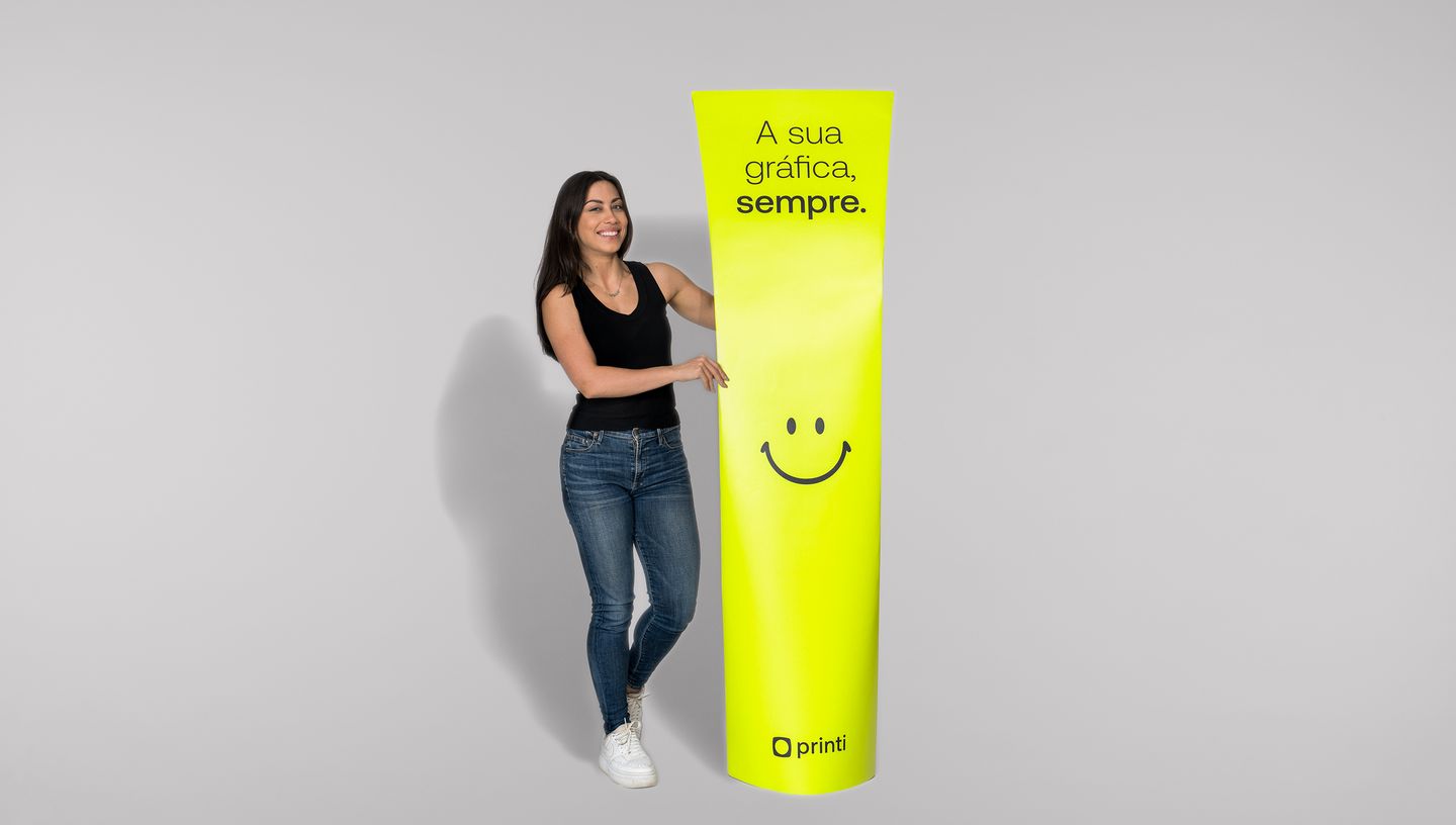 Mulher branca de cabelos lisos com regata preta, calça jeans skinny e tênis branco segurando totem banana com smile na cor verde-limão com fundo cinza