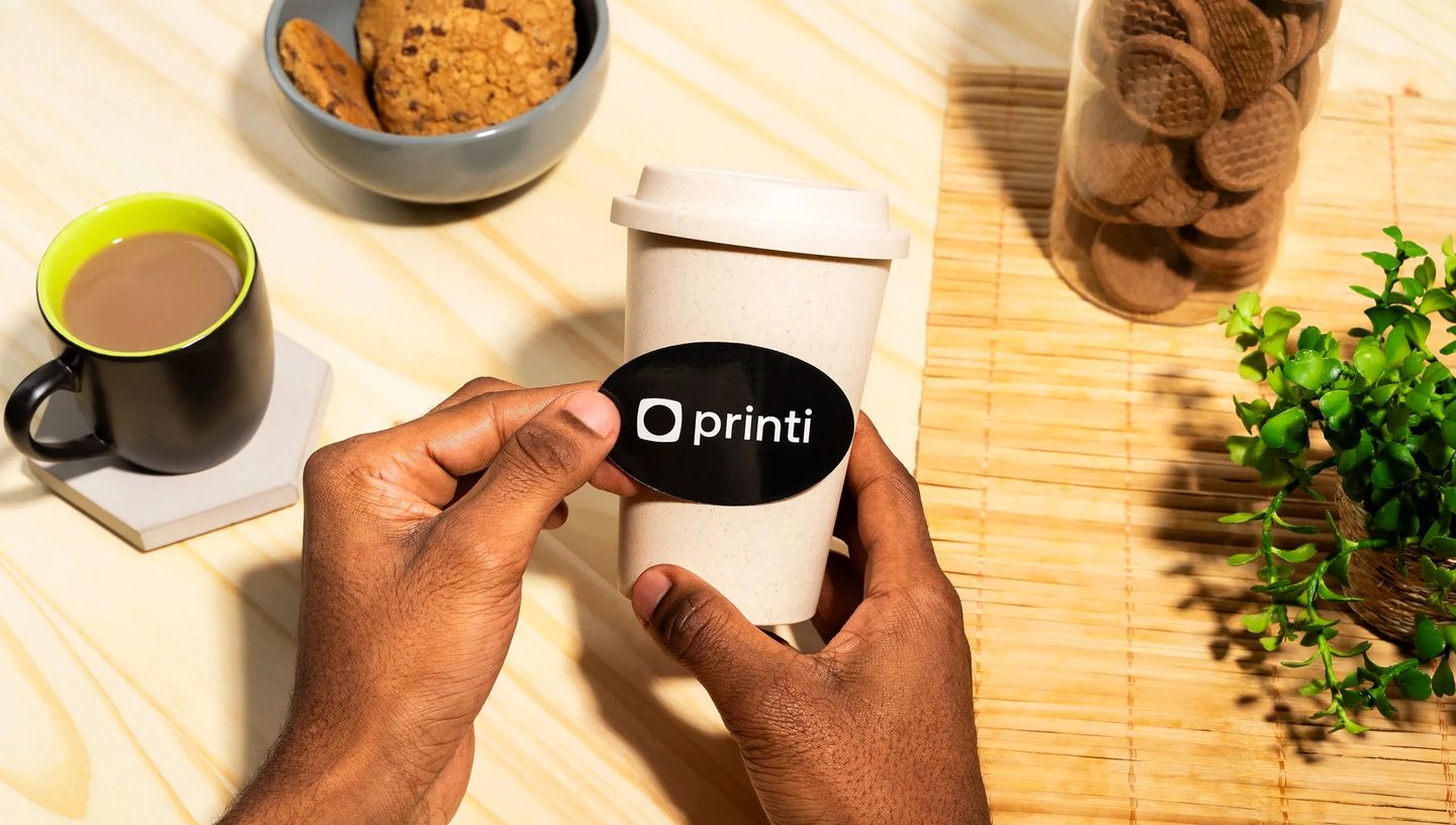 Adesivo oval personalizado preto com logotipo Printi aplicado sobre copo térmico sustentável.