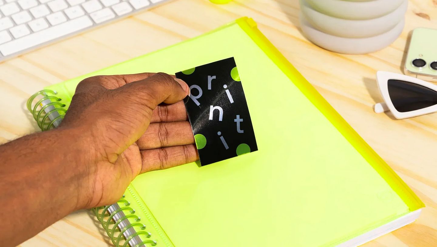 Mão segurando adesivo retangular personalizado da Printi sobre caderno verde neon em mesa de escritório, demonstrando aplicação prática do material.