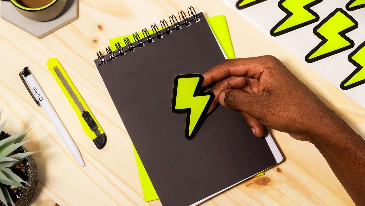 Mão aplicando adesivo personalizado de raio em caderno preto, demonstrando a aderência e o uso de papelaria personalizada em escritório.