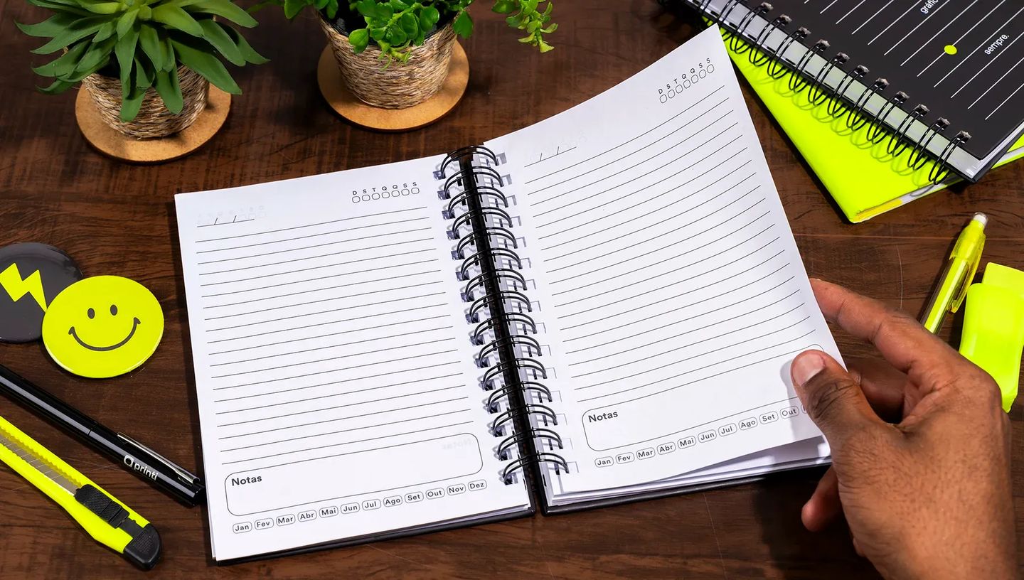 Agenda permanente personalizada da Printi sobre mesa de madeira ao lado de notebook e café, demonstrando o uso profissional e acadêmico do produto em ambiente de escritório.