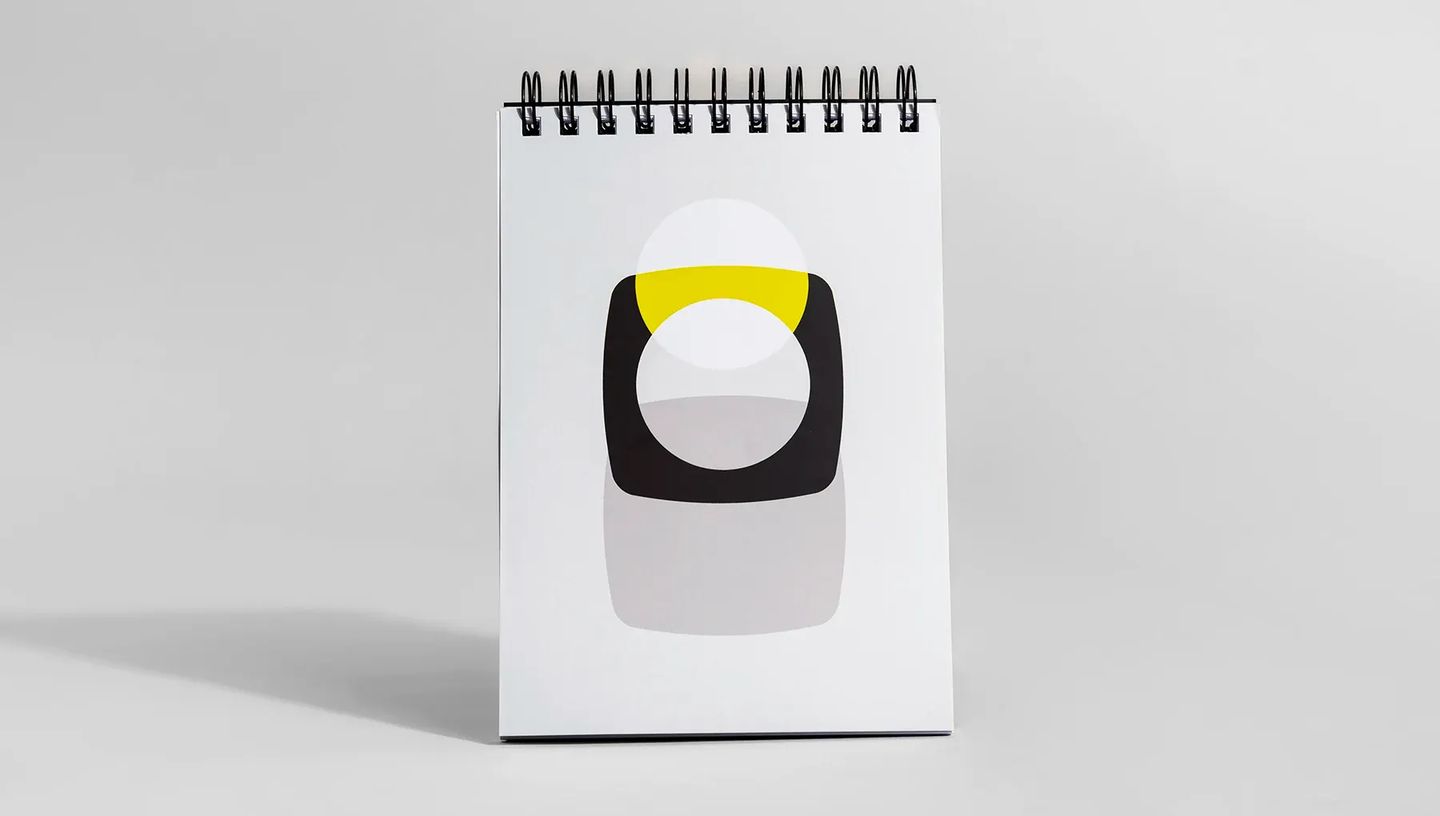 Bloco de notas com capa wire-o posicionado verticalmente, apresentando design minimalista com formas geométricas em preto e amarelo sobre fundo cinza claro.