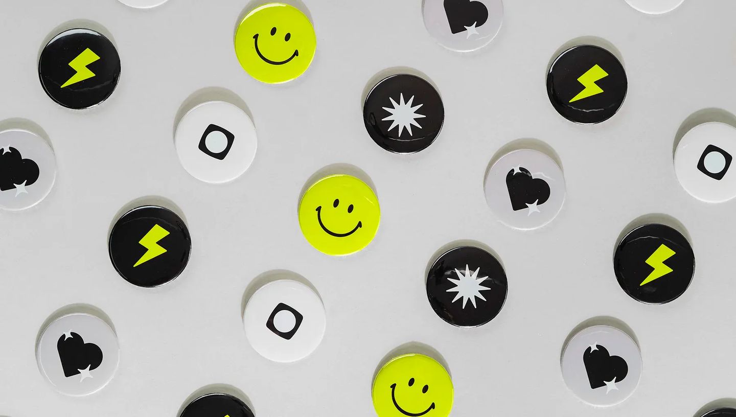 Composição de diversos bottons personalizados redondos com logotipos e ícones variados em preto, branco e amarelo.