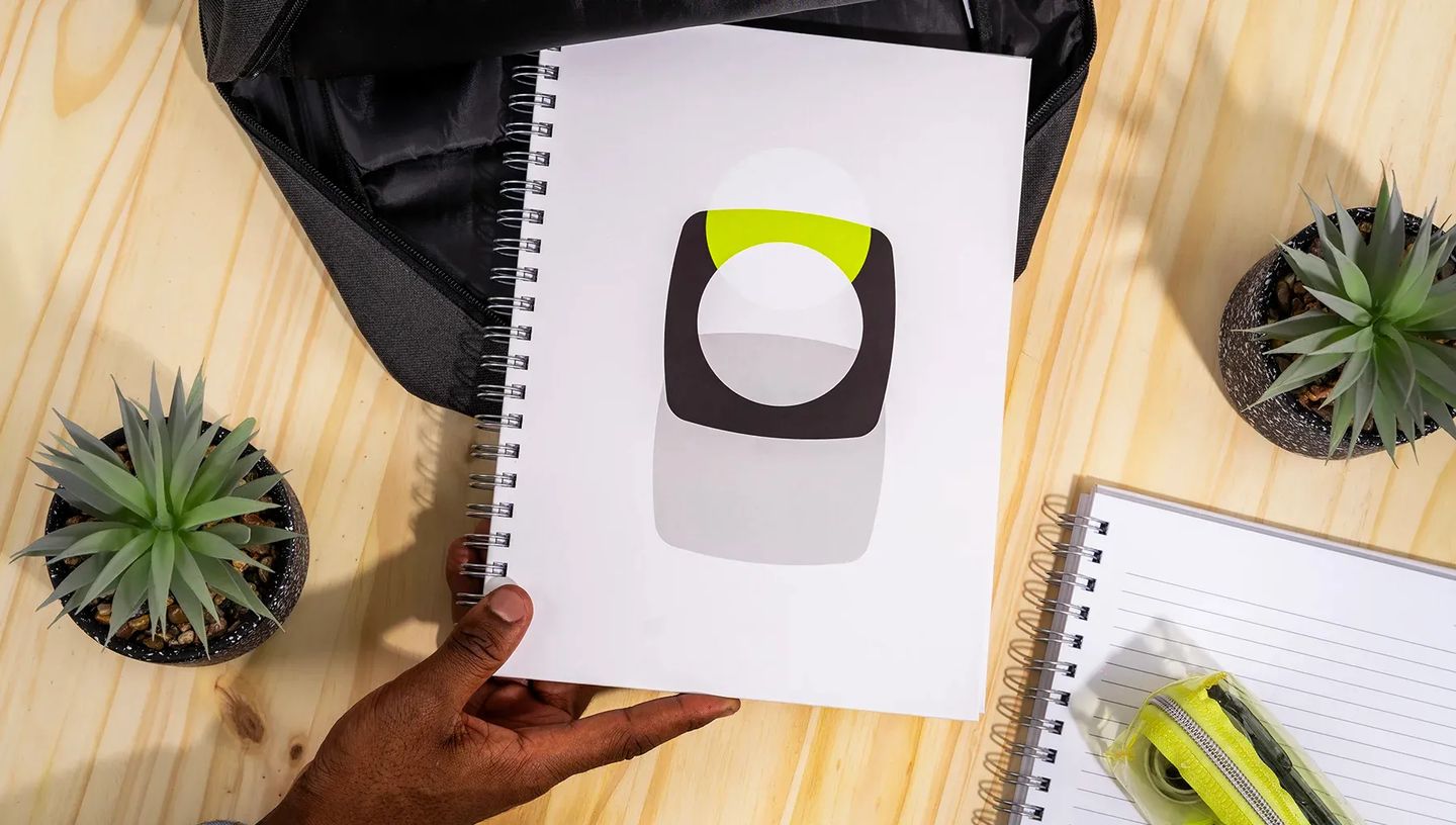 Pessoa segurando caderno personalizado da Printi em mesa de madeira ao lado de mochila e acessórios de papelaria, destacando o uso acadêmico e profissional.