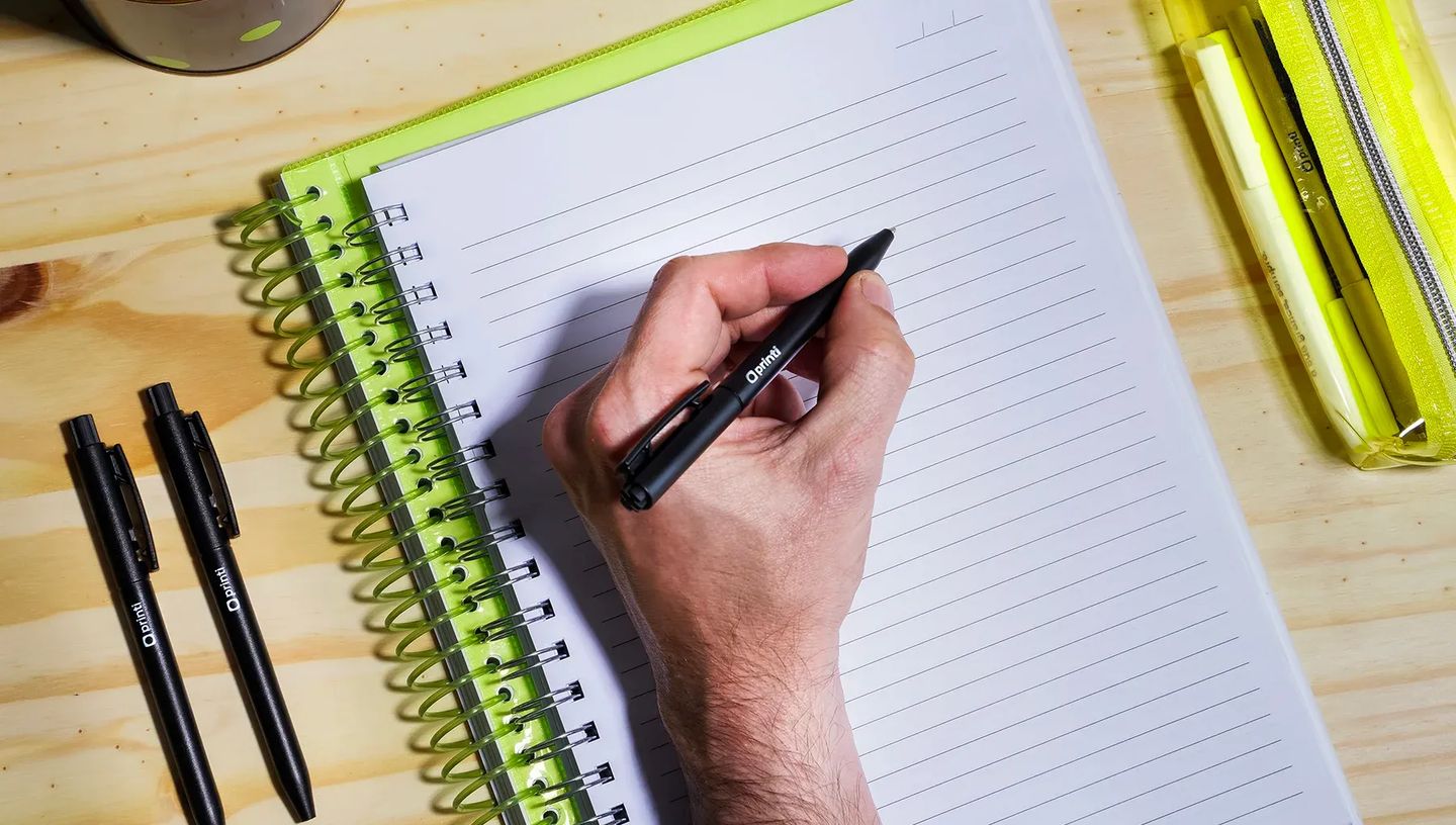 Close-up de duas canetas plásticas personalizadas da Printi, nas cores laranja e verde limão, apoiadas sobre um caderno de capa dura preto. A imagem destaca o clipe de bolso cinza e a ponteira prateada das canetas em um ambiente de escritório, enfatizando