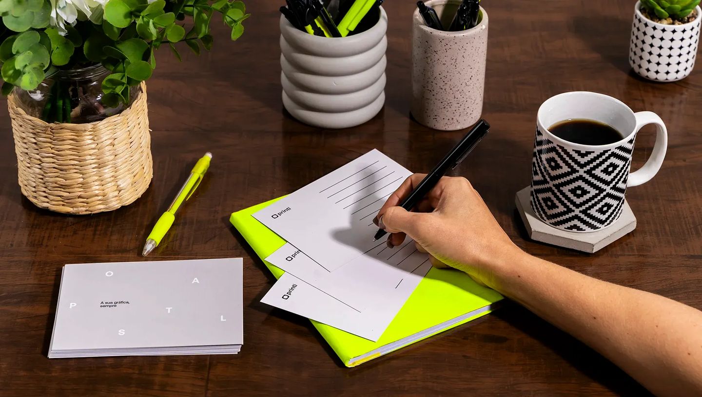 Pessoa escrevendo em cartão postal personalizado da Printi sobre mesa de escritório com café e acessórios de papelaria.