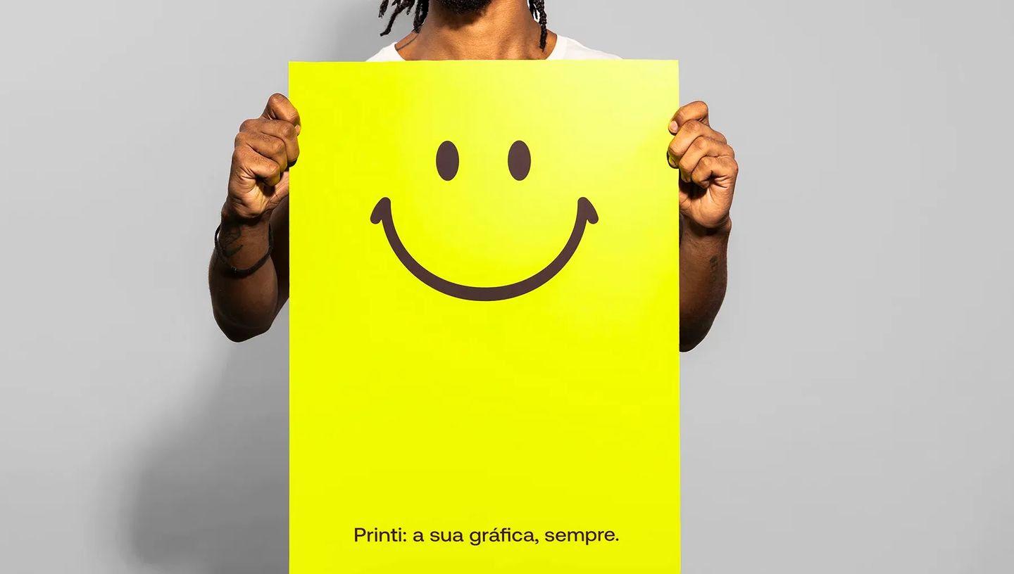 Homem segurando cartaz personalizado grande em amarelo neon com ícone de sorriso e a frase "Printi: a sua gráfica, sempre", demonstrando impacto visual.
