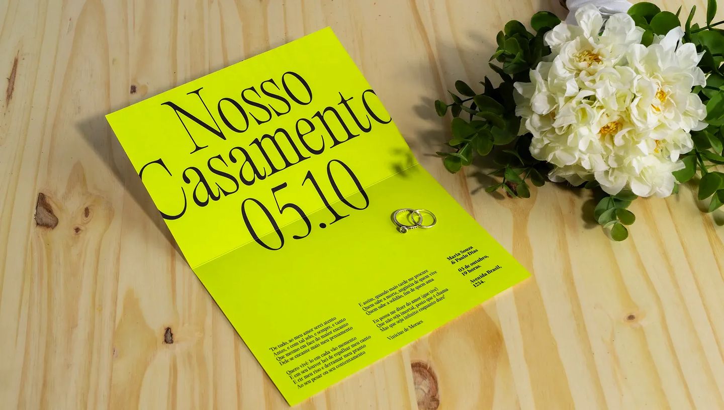 Convite de casamento moderno em papel verde-limão neon aberto sobre mesa de madeira, exibindo poema de Vinicius de Moraes e detalhes do evento ao lado de duas alianças e um buquê de flores brancas.