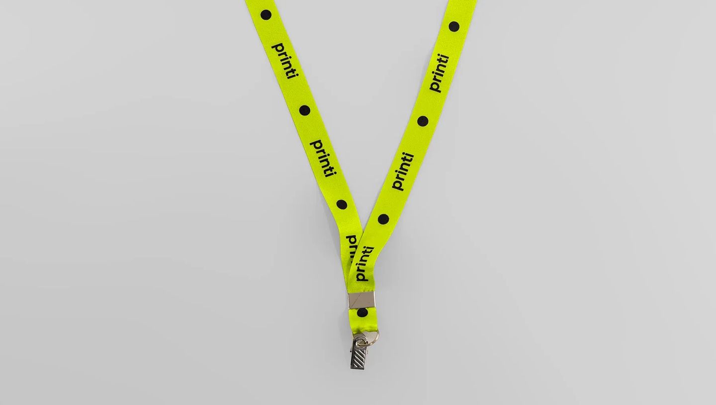 Cordão para crachá na cor verde, dobrado em forma de 'V', sobre uma superfície cinza neutra. A fita tem a marca 'Printi' impressa em preto com pontos pretos