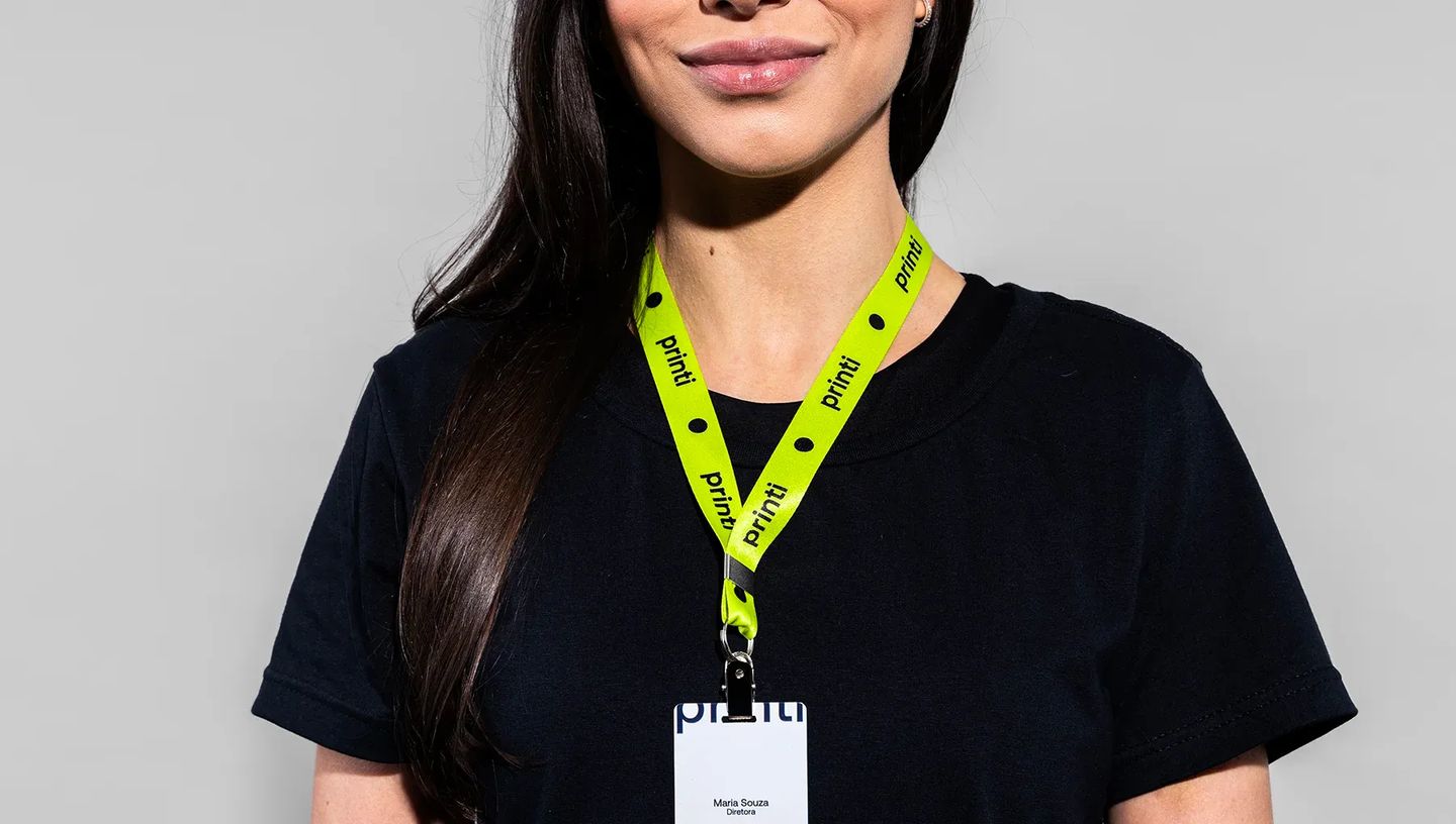 Mulher sorrindo, vestindo uma camiseta preta e um cordão para crachá verde neon. O cordão exibe o logo preto da Printi repetido e está preso a um crachá de identificação. O fundo é cinza claro.