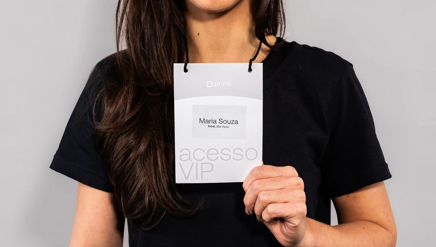 Mulher vestindo camiseta preta segura uma credencial de identificação personalizada com cordão preto. O crachá exibe o logotipo da Printi, o nome "Maria Souza" e a inscrição "acesso VIP" em destaque.