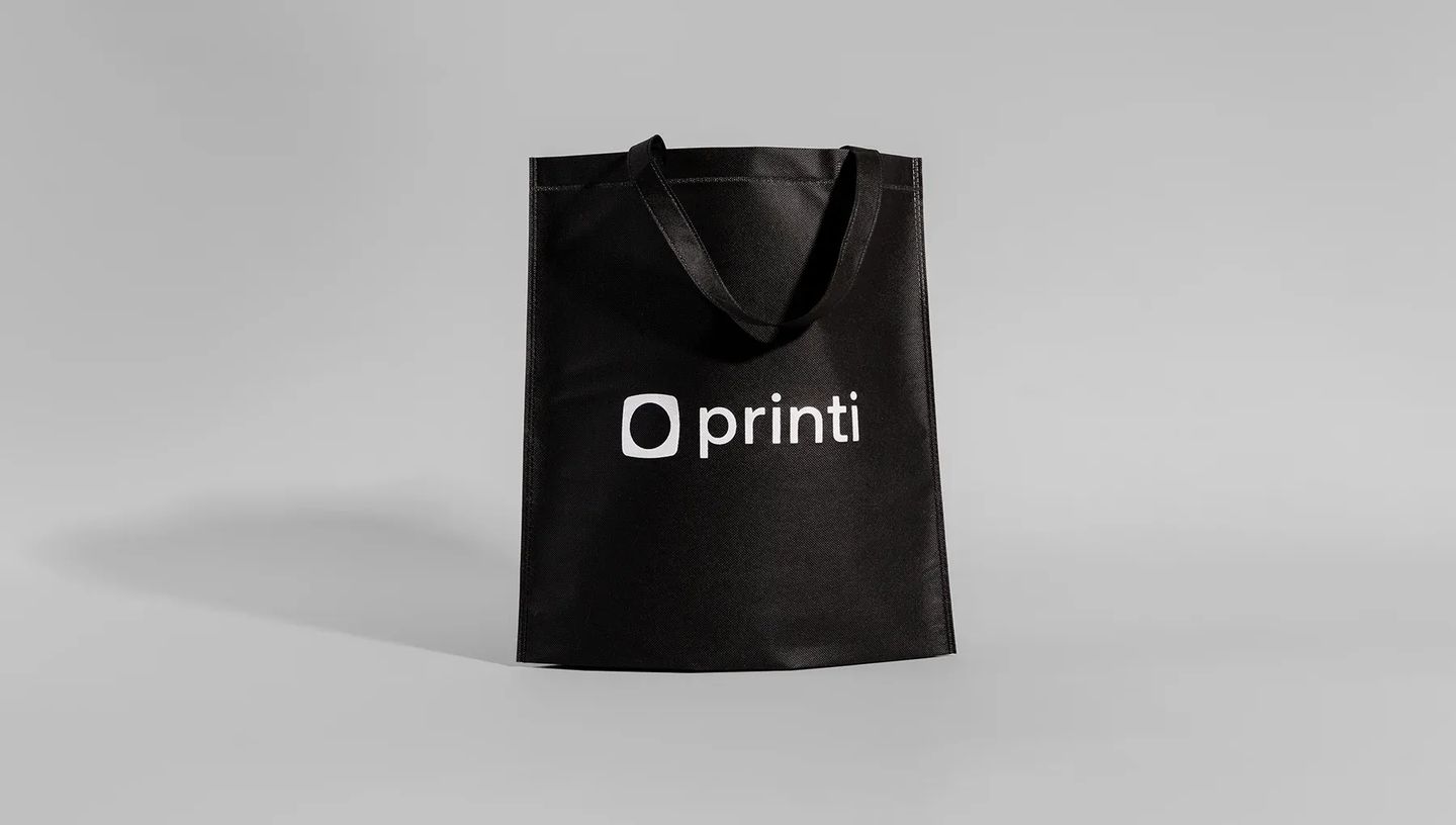 Ecobag personalizada de TNT preta com o logo da Printi centralizado, em pé contra um fundo cinza.