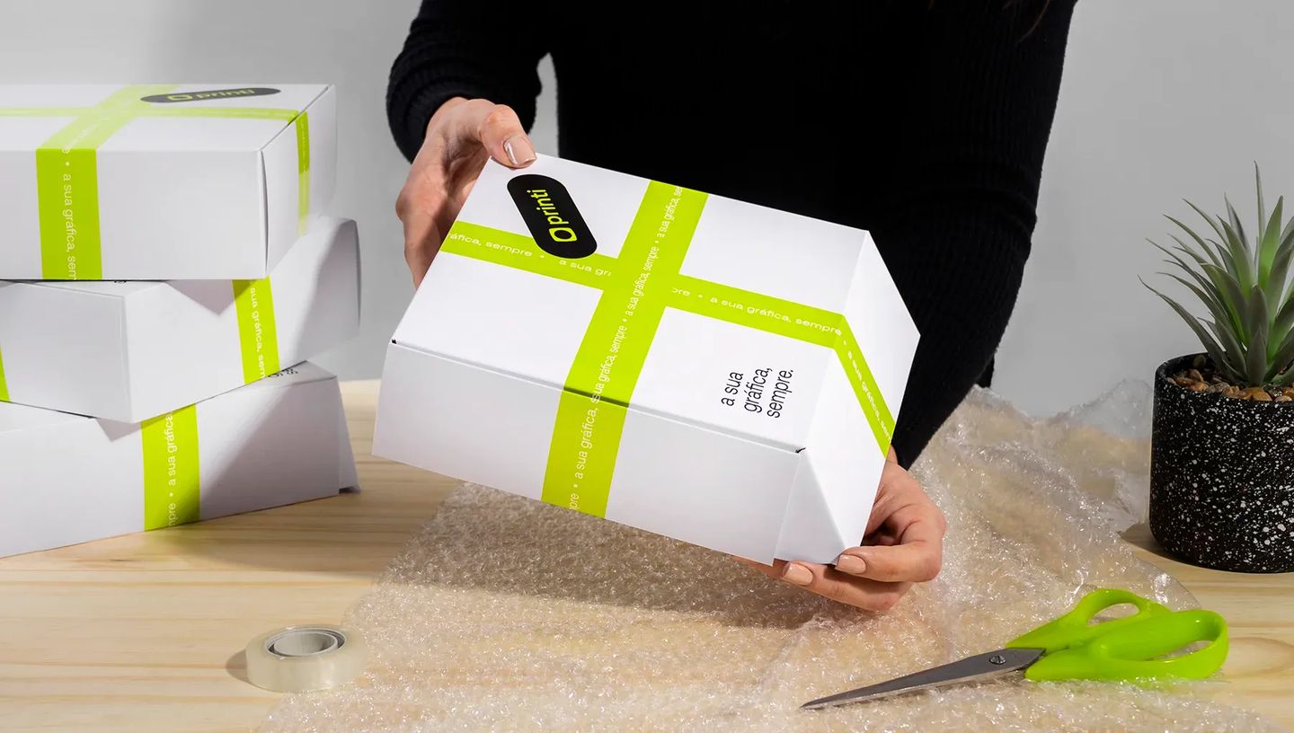 Pessoa manuseando caixa basculante personalizada da Printi em mesa de expedição com plástico bolha, evidenciando o uso prático para envios.