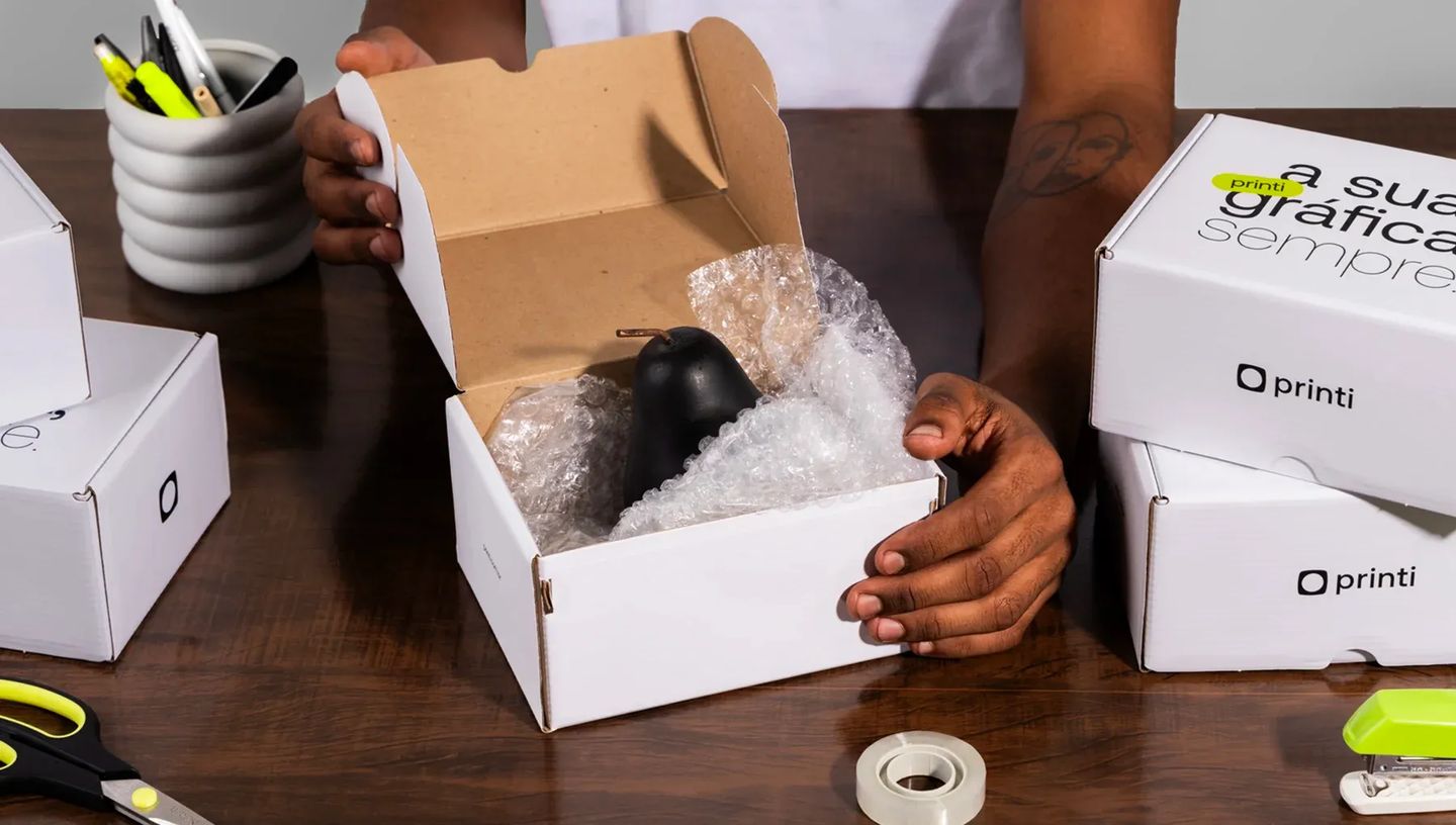 Pessoa embalando objeto em caixa de correio personalizada da Printi com plástico bolha, destacando a proteção e segurança para logística de e-commerce.