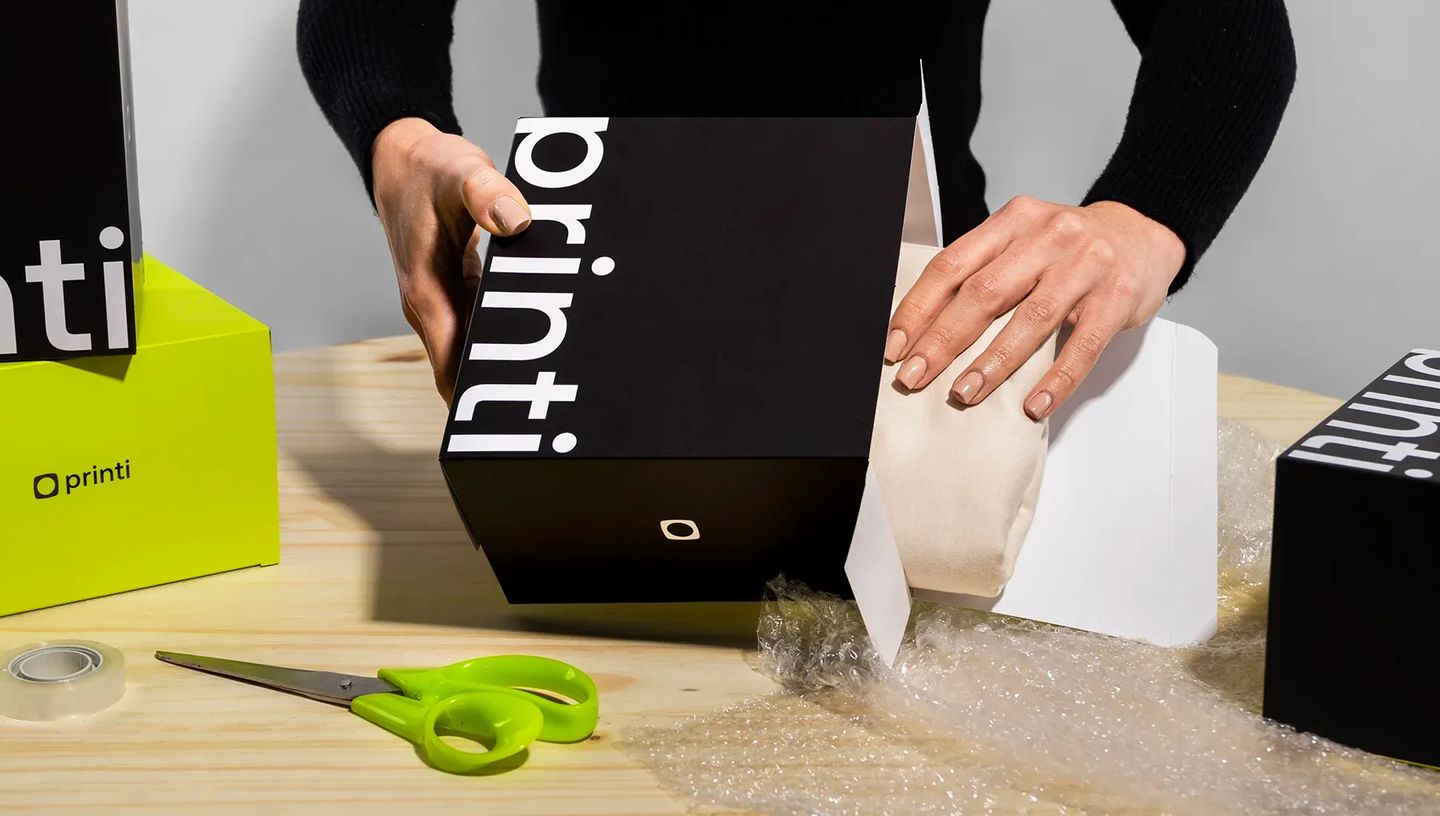 Pessoa fazendo unboxing e colocando produto em embalagem personalizada da Printi. Sobre uma mesa de madeira, há uma caixa preta com logotipo branco, plástico bolha, tesoura e outras caixas institucionais em verde e preto.