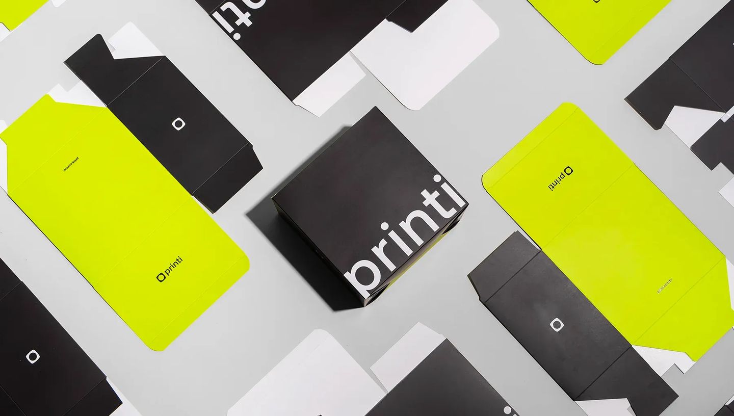 Foto com vista superior de diversas embalagens de papel desmontadas e uma montada da marca Printi. O design utiliza um contraste forte entre preto, verde-limão vibrante e branco. A caixa central, já montada, é preta com o logotipo "printi" em letras branc