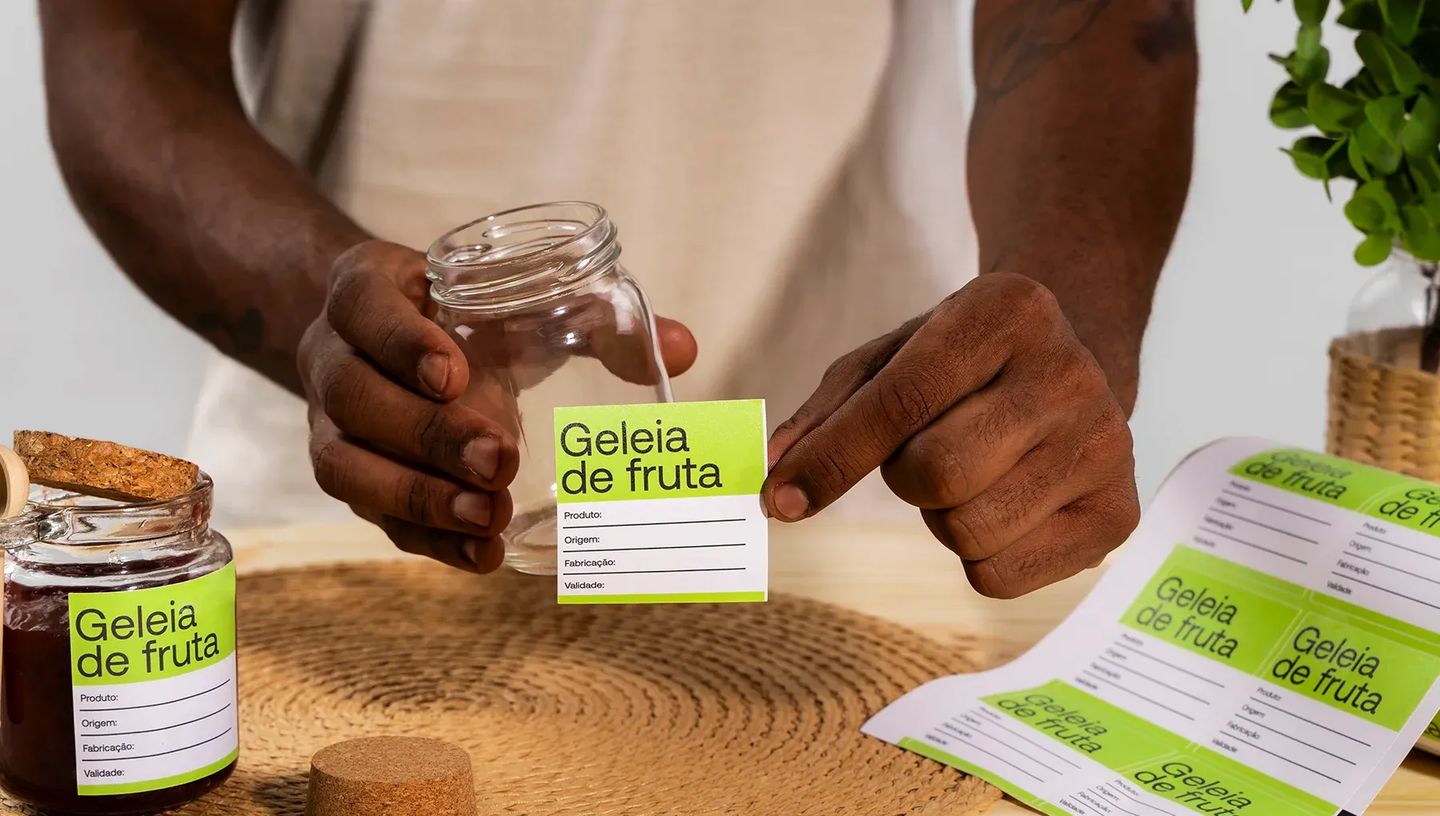 Pessoa aplicando etiqueta personalizada para geleia de fruta em pote de vidro, destacando o uso para rotulagem de alimentos e informações de validade.