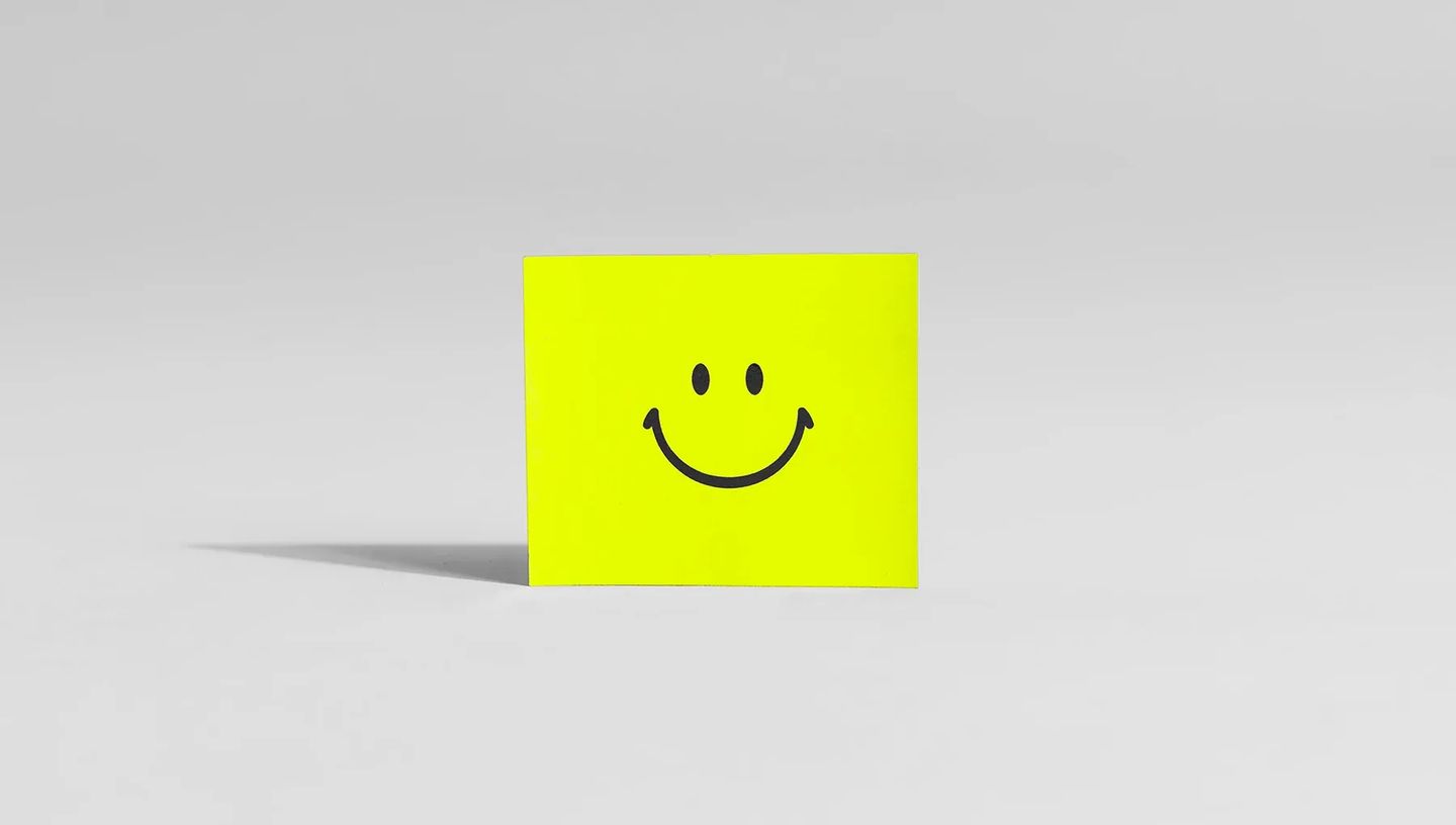 Ímã de geladeira quadrado na cor amarelo neon com estampa de emoji de carinha sorridente preta, centralizado em um fundo branco minimalista com sombra projetada lateralmente.