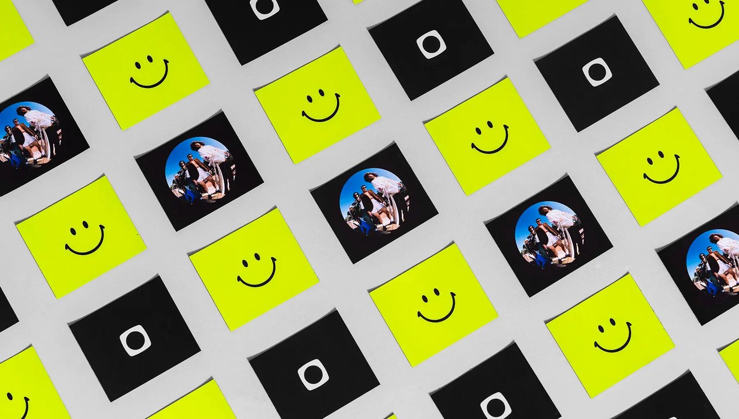 Vários modelos de ímã de geladeira quadrado espalhados, alternando entre estampas de emoji de carinha sorridente amarela, fotos de pessoas e um logotipo branco em fundo preto.