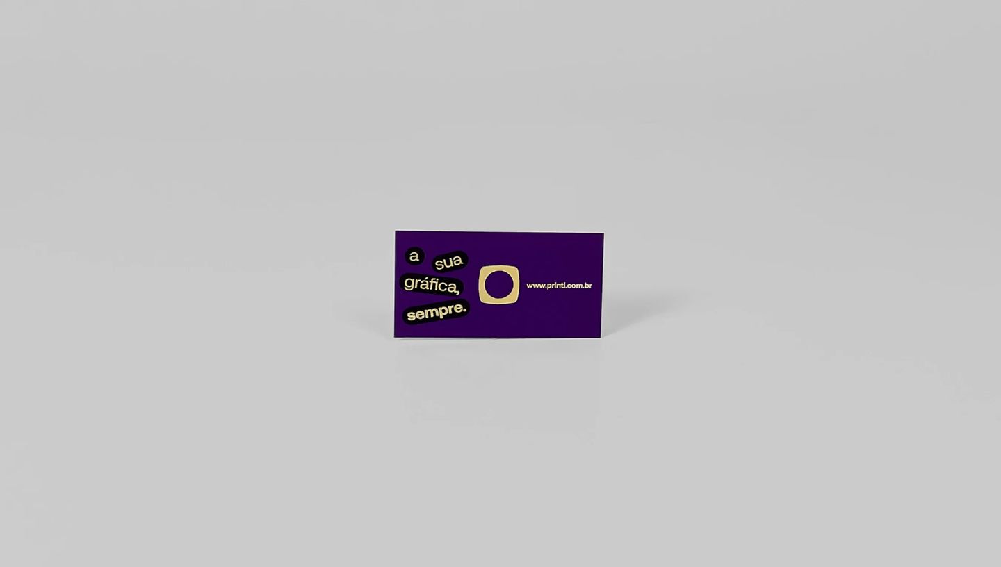 Cartão de visita roxo com acabamento em hot stamping dourado, com a frase “a sua gráfica, sempre” e logotipo Printi, posicionado sobre fundo cinza claro.