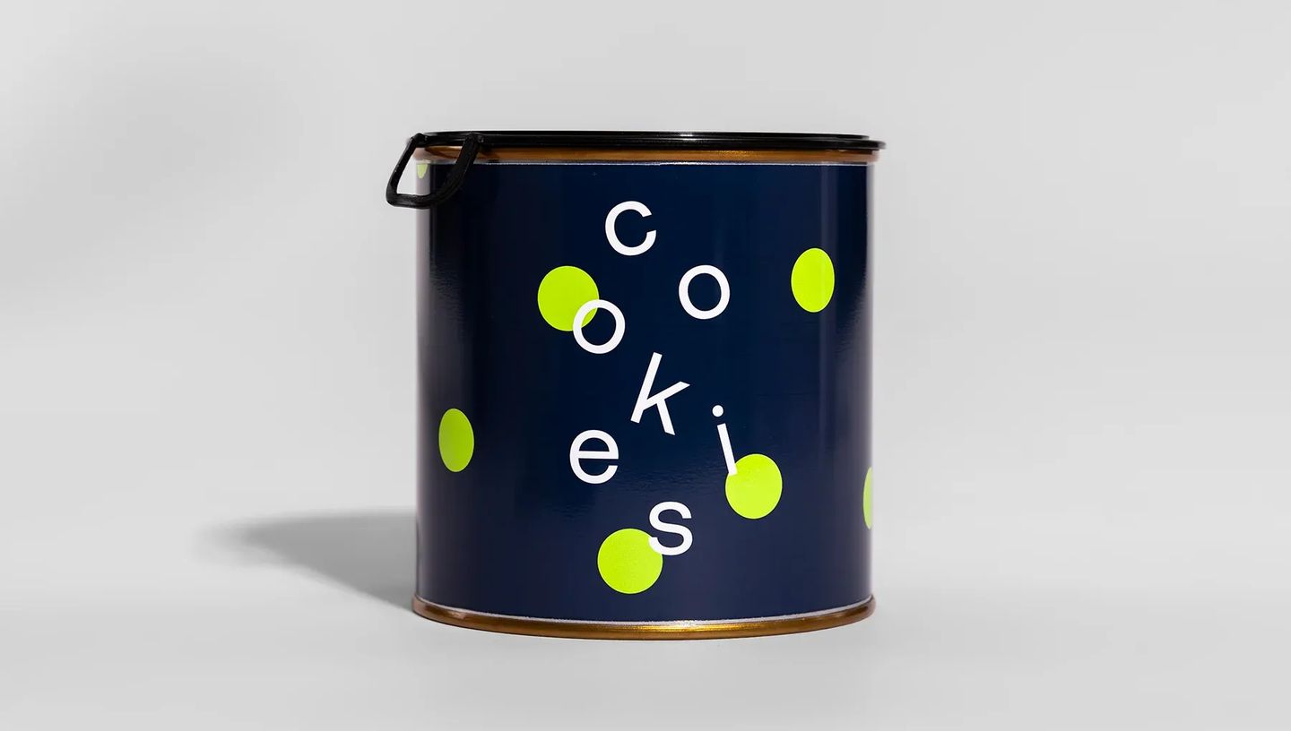 Lata de metal cilíndrica com design moderno na cor azul-marinho, estampa de bolinhas verdes e a palavra cookies, com tampa preta fechada em fundo cinza claro.