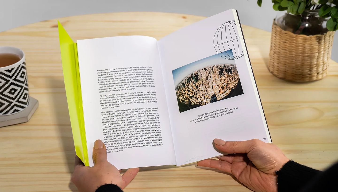 Visão interna de livro personalizado da Printi aberto em mesa de madeira, exibindo diagramação profissional com textos e fotografia de paisagem urbana.