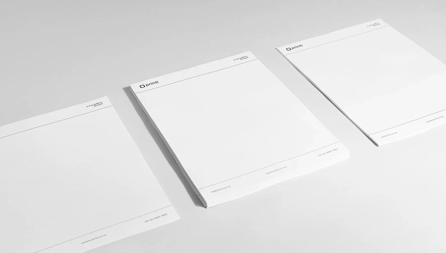 Três blocos de papel timbrado personalizado da Printi dispostos em perspectiva sobre fundo branco. Cada bloco exibe o logotipo no topo e o slogan institucional, destacando a produção em escala de papelaria corporativa profissional.