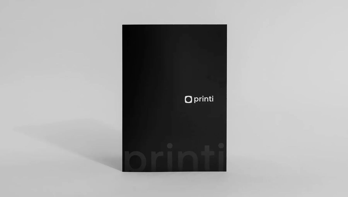 Pasta personalizada da Printi em papel preto fosco com acabamento premium. O design apresenta o logotipo centralizado em branco e uma marca d'água discreta da marca na parte inferior, sobre um fundo neutro.