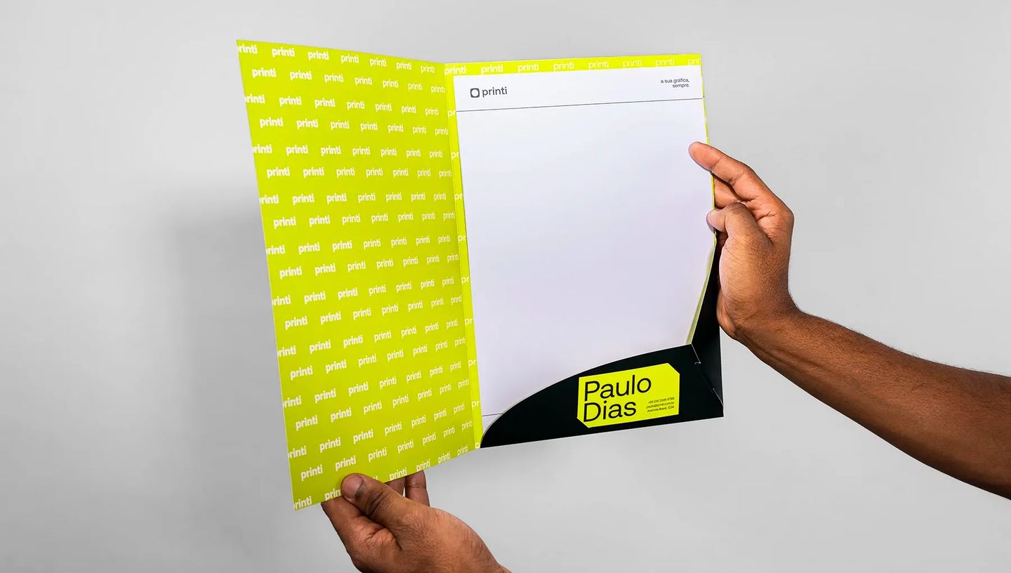Mãos segurando pasta personalizada aberta com bolsa interna preta e porta-cartão. O interior é decorado com padrão de logotipos da Printi em fundo verde-limão e contém folhas brancas de papel timbrado.