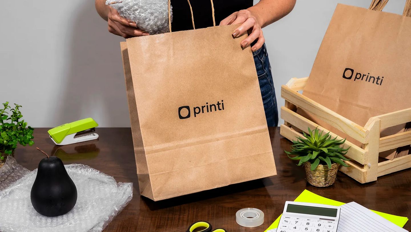 Sacola de papel kraft personalizada da Printi sendo usada para embalar produtos com plástico bolha em uma mesa de trabalho organizada. Ideal para e-commerce e lojas físicas.