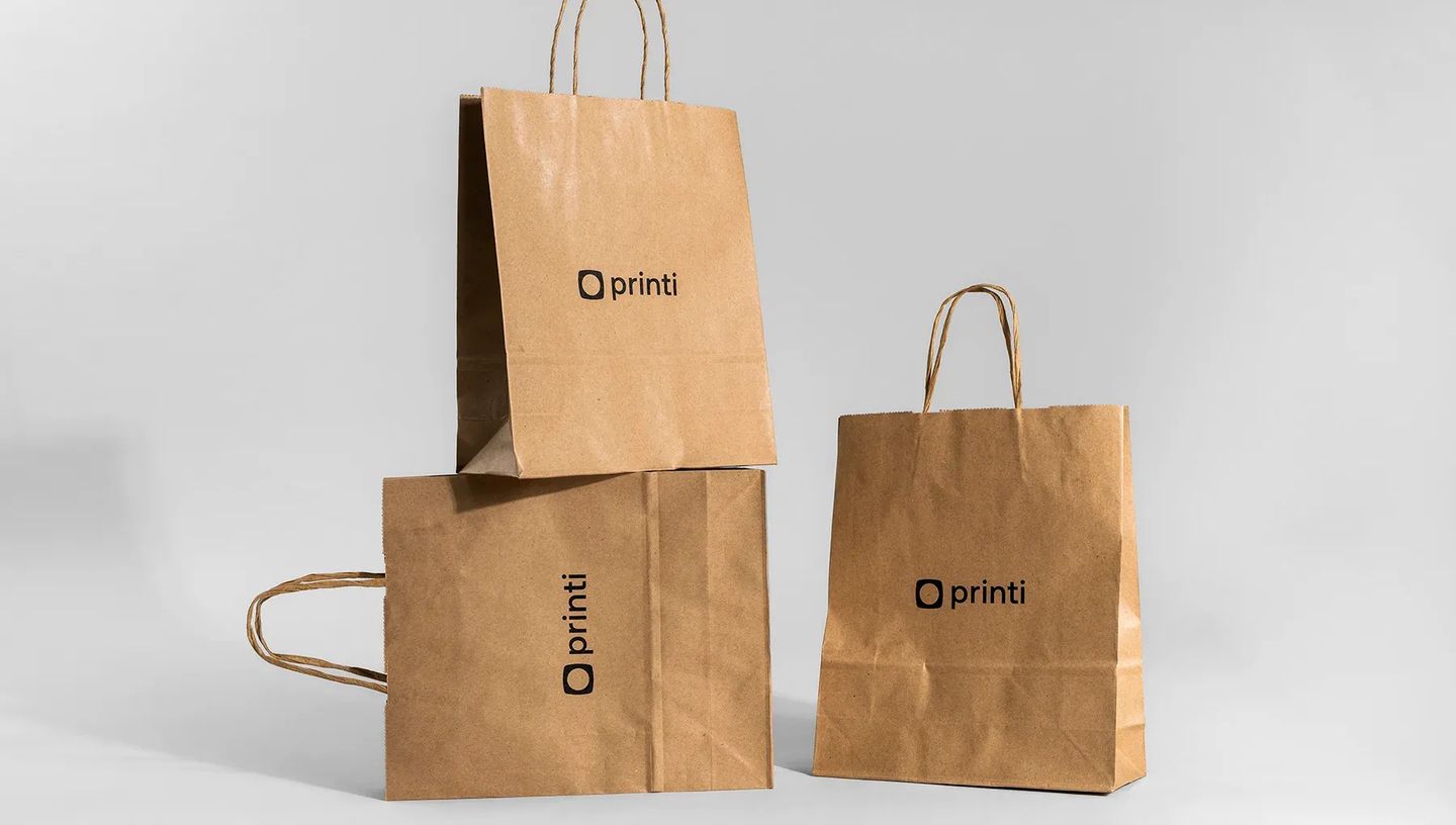 Três sacolas de papel kraft personalizadas empilhadas em fundo cinza claro, destacando o design, a resistência e o tamanho da embalagem corporativa.