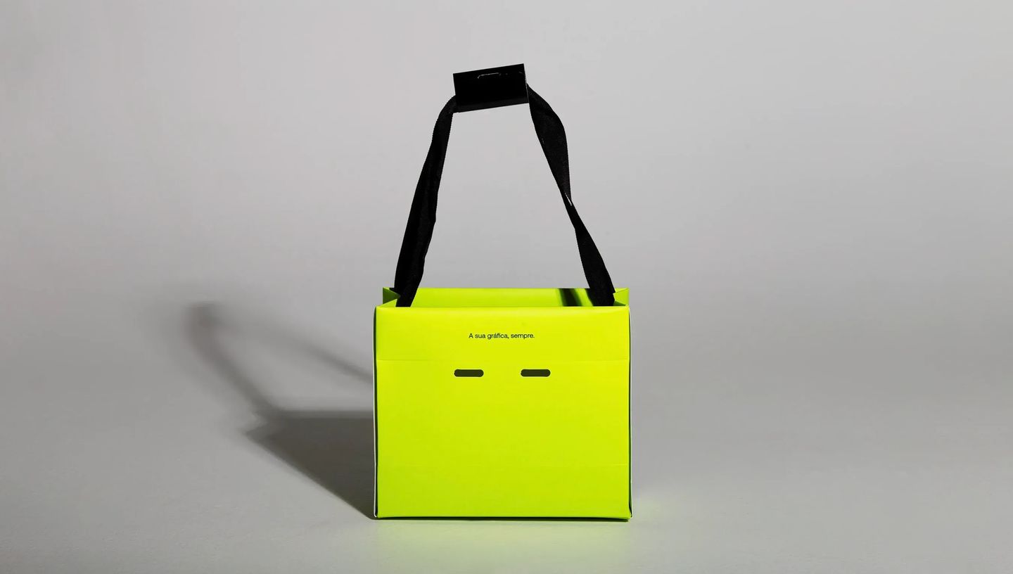 Sacola de papel personalizada modelo teepee em verde neon com estampa de ícone sorridente (smile) e alças pretas em fundo cinza neutro.