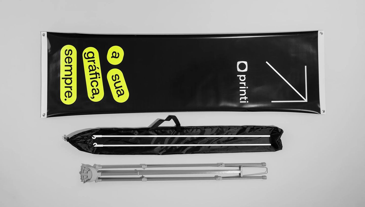 Banner X personalizado da Printi em fundo preto com elementos gráficos em verde-limão e a frase “a sua gráfica, sempre”, acompanhado de estrutura metálica desmontável e bolsa de transporte, utilizado como material de comunicação visual para feiras, evento