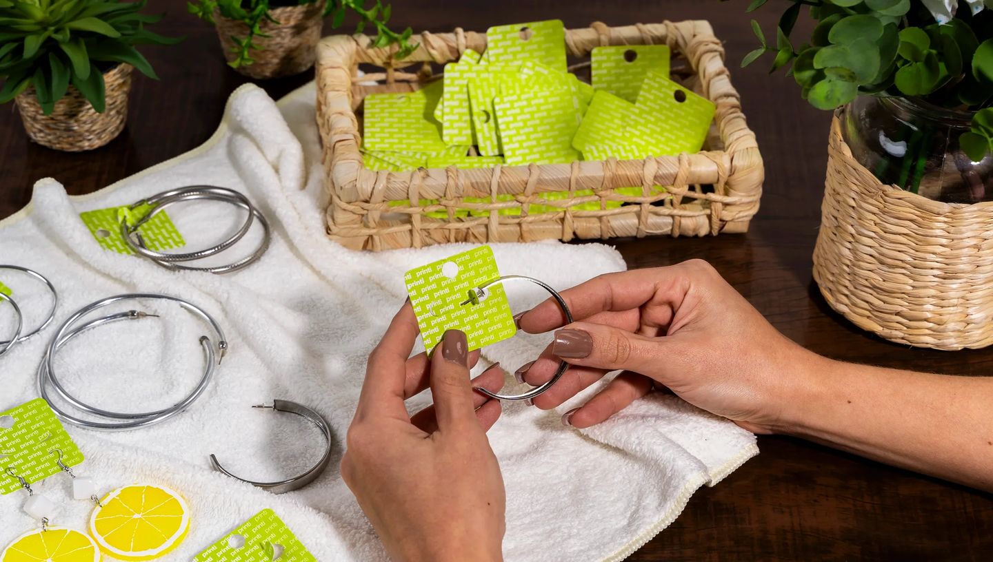 Mãos de uma pessoa branca encaixando brinco de argola em cartela para bijuteria personalizada da Printi, com outras argolas de prata organizadas sobre uma toalha branca.