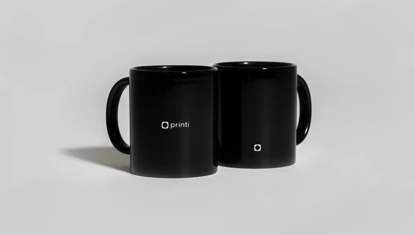 Duas canecas pretas personalizadas da Printi em cerâmica sobre fundo neutro, com logotipo branco