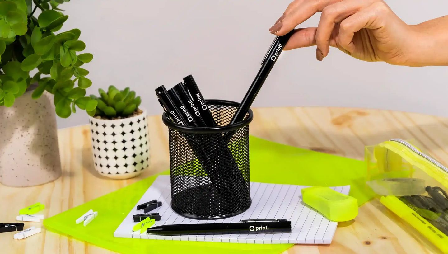 Mão retirando caneta metálica personalizada com logo Printi de um porta-canetas preto sobre mesa de madeira, cercada por itens de papelaria como marca-texto, clips, caderno pautado e estojo transparente, além de plantas decorativas, representando uma cane