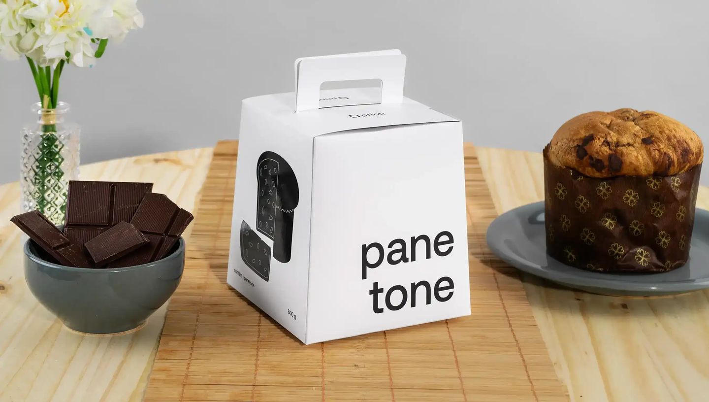 Embalagem personalizada para panetone com alça em caixa branca sobre mesa de madeira, acompanhada de panetone em prato e pedaços de chocolate em tigela