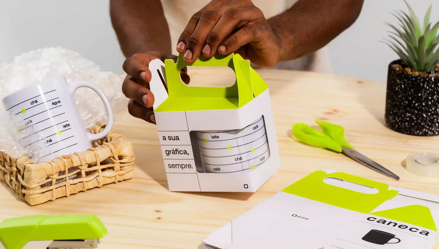 Embalagem personalizada para caneca sendo montada em mesa de trabalho, com design em branco e verde neon da Printi, ideal para presentear, proteger e valorizar canecas personalizadas em brindes ou vendas.