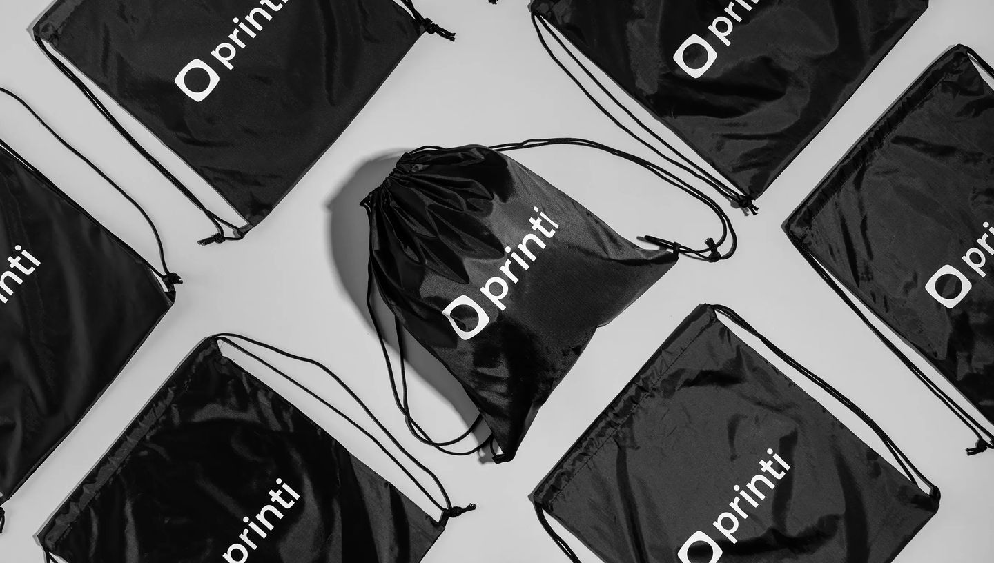 Várias mochilas saco personalizadas na cor preta, com estampa do logo da Printi, organizadas em fundo claro.